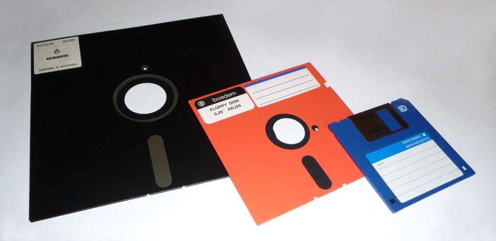 Three floppy disks are displayed: a large black 8-inch disk, a middle-sized red 5.25-inch disk, and a smaller blue 3.5-inch disk. Each has a central circular cutout and a label area, showing the evolution of floppy disk sizes.