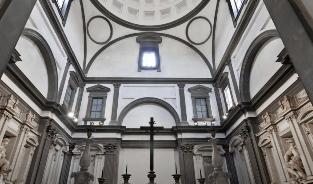 Interior of a chapel with a black cross in the center, surrounded by classical architecture. The walls are adorned with stone columns and statues. Large windows at the top let in natural light, highlighting the geometric patterns on the ceiling.