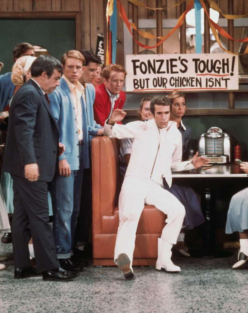 A group of people stand around a diner booth where a person in a white suit and boots strikes a pose. Above, a banner reads, "FONZIE'S TOUGH! BUT OUR CHICKEN ISN'T." A jukebox is visible in the background.