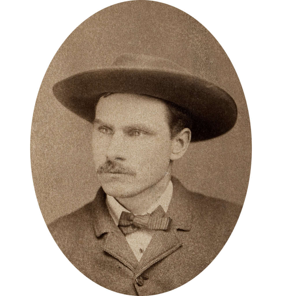 A sepia-toned vintage portrait of a man wearing a wide-brimmed hat and a suit with a bow tie. He has a mustache and is looking off to the side with a serious expression. The image has an oval frame.