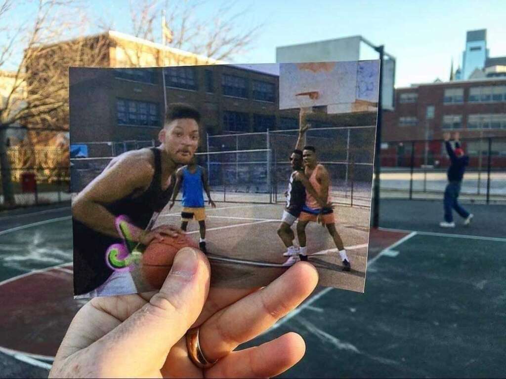 A hand holds a photo of people playing basketball, perfectly aligning with an outdoor court scene in the background. The old photo shows players in action, while the court behind it is mostly empty, except for a person walking.