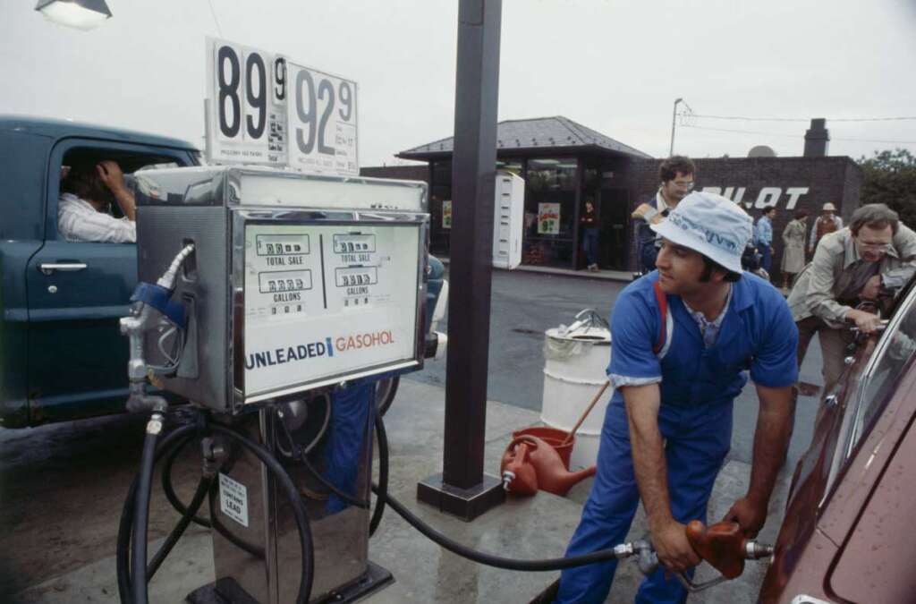 A man in a blue jumpsuit refuels a car at a gas station. Gas prices are displayed as 89.9 and 92.9 cents. A person is seated in a truck in the background, while others are seen around the station.