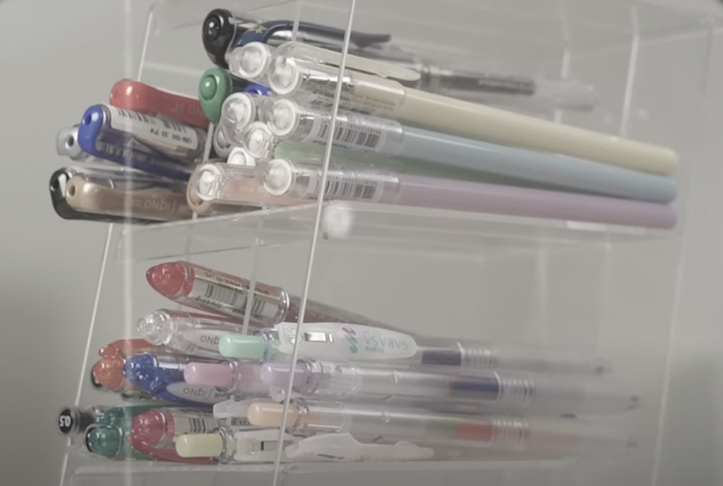 A clear plastic organizer holds various colorful pens and pencils, including pastel-colored mechanical pencils and multicolored pens with transparent barrels. The organizer has multiple compartments, displaying the items neatly.