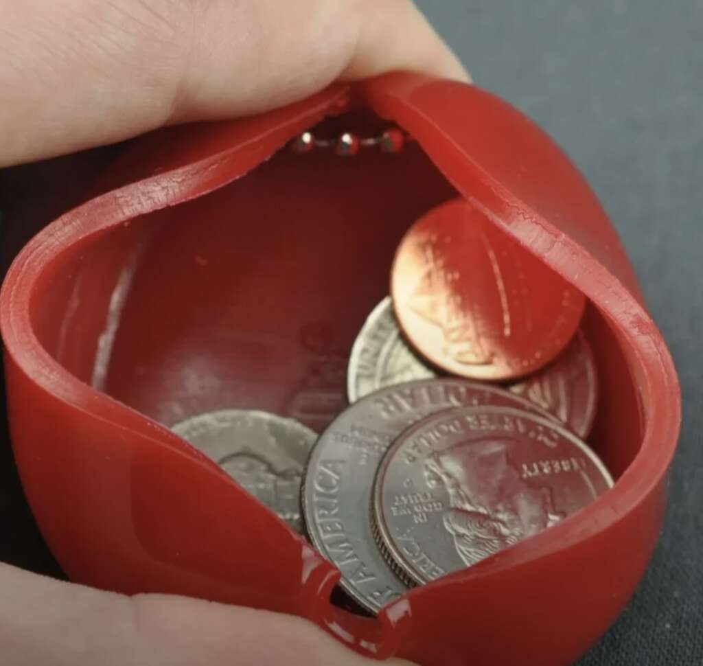 A close-up of a red, clamshell-style coin purse, slightly open, revealing various coins inside, including quarters and pennies, on a dark surface. A hand is gently holding the purse open.