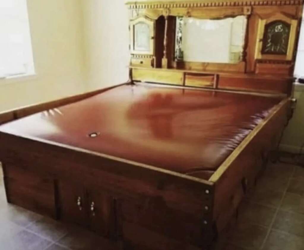 A vintage wooden waterbed with an ornate mirrored headboard and built-in storage. The bed is filled with water, creating a smooth, wavy appearance on the surface. The room has tiled flooring and a window to the left.