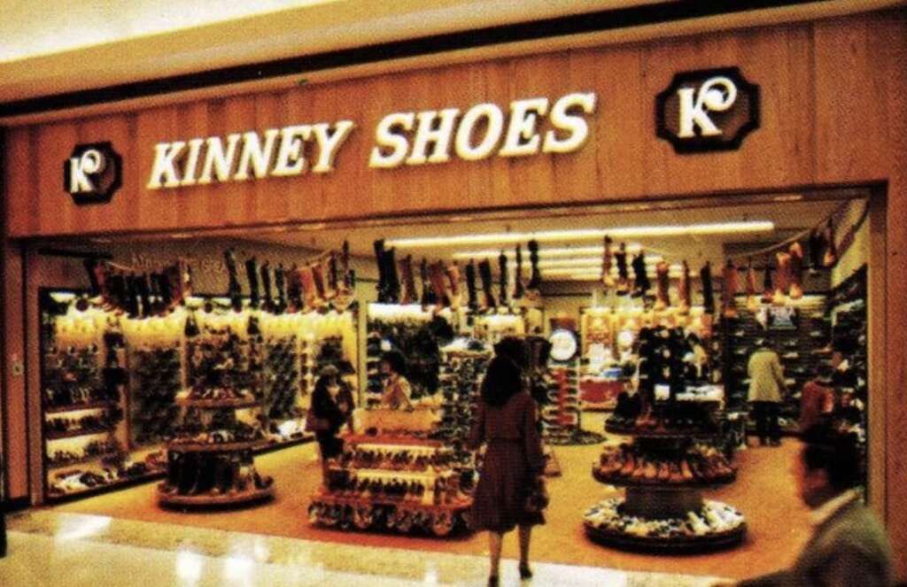 Vintage photo of a Kinney Shoes store displaying a variety of footwear. A woman is walking towards the entrance. The interior is brightly lit with neatly arranged shoes on shelves and displays.