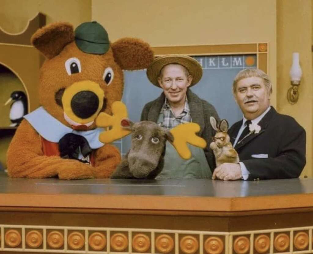 A group of characters stand behind a counter. A person in a bear costume, a large puppet of a moose, a man in overalls and straw hat, a small rabbit puppet, and a man in a suit with a flower pinned to his lapel are featured, with a penguin statue nearby.