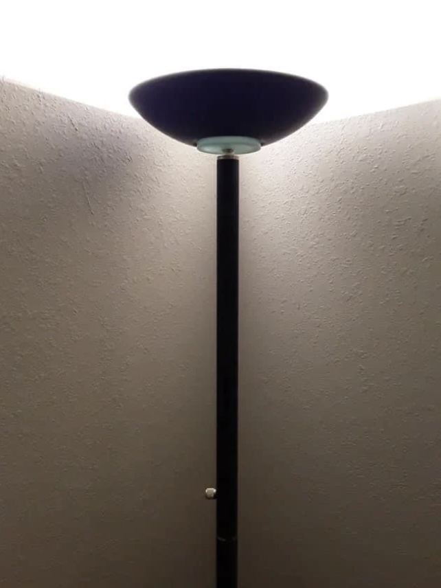 A tall, black floor lamp stands in the corner of a room, casting a bright light upward against two beige walls. The base and pole of the lamp are visible, with a glowing lampshade at the top.