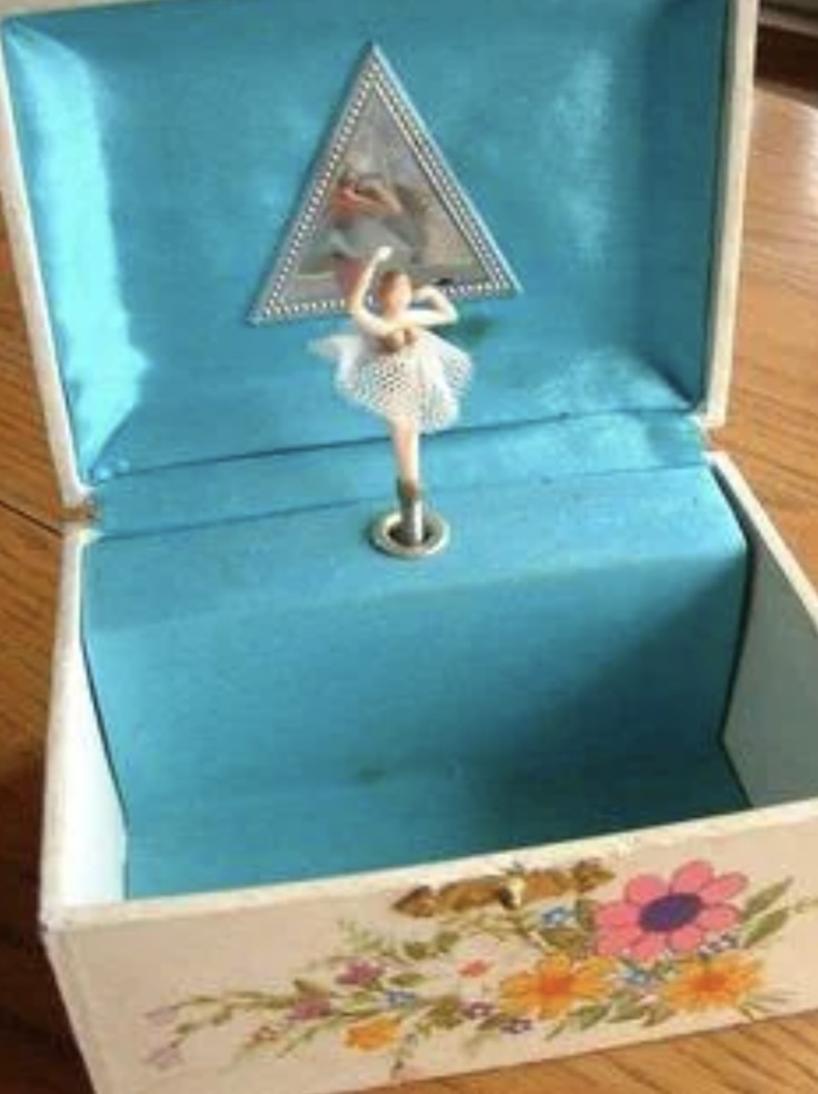 An open music box featuring a small ballerina figurine wearing a white tutu, positioned in front of a triangular mirror. The box is lined with light blue fabric, and the exterior displays colorful floral designs.