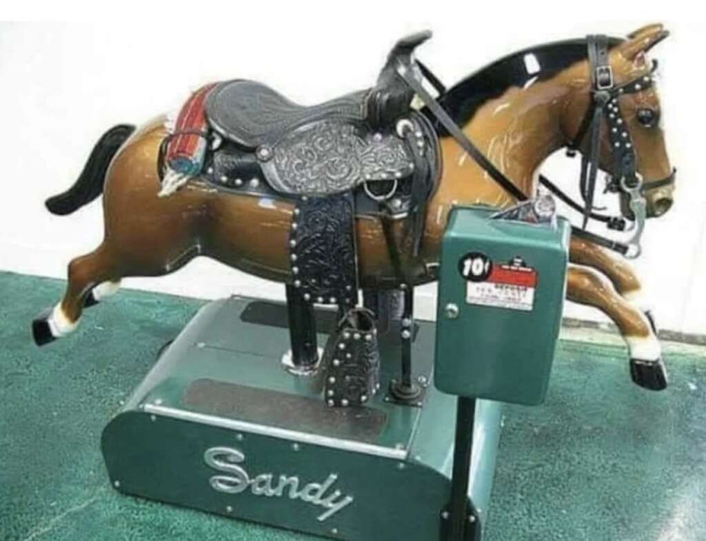 A vintage mechanical horse ride named "Sandy" with a brown body, saddle, and bridle. It is mounted on a green base with a coin-operated control box labeled with "101." The setup sits on a green floor.