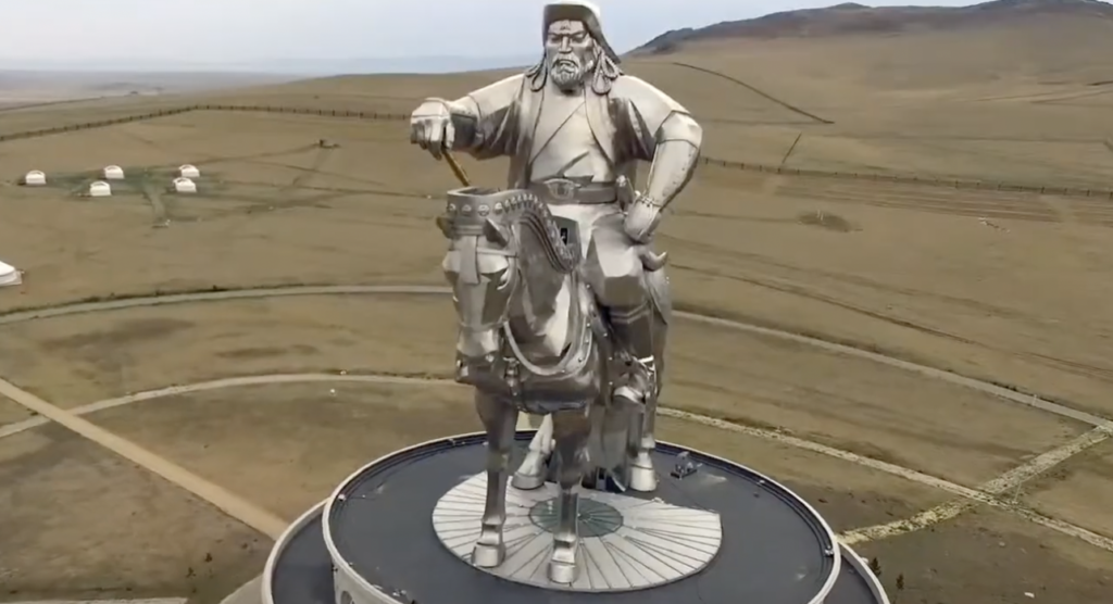 Aerial view of a large silver statue of a man on horseback, holding a golden whip, on a circular platform. The statue overlooks a vast, flat landscape with a few scattered white tents.