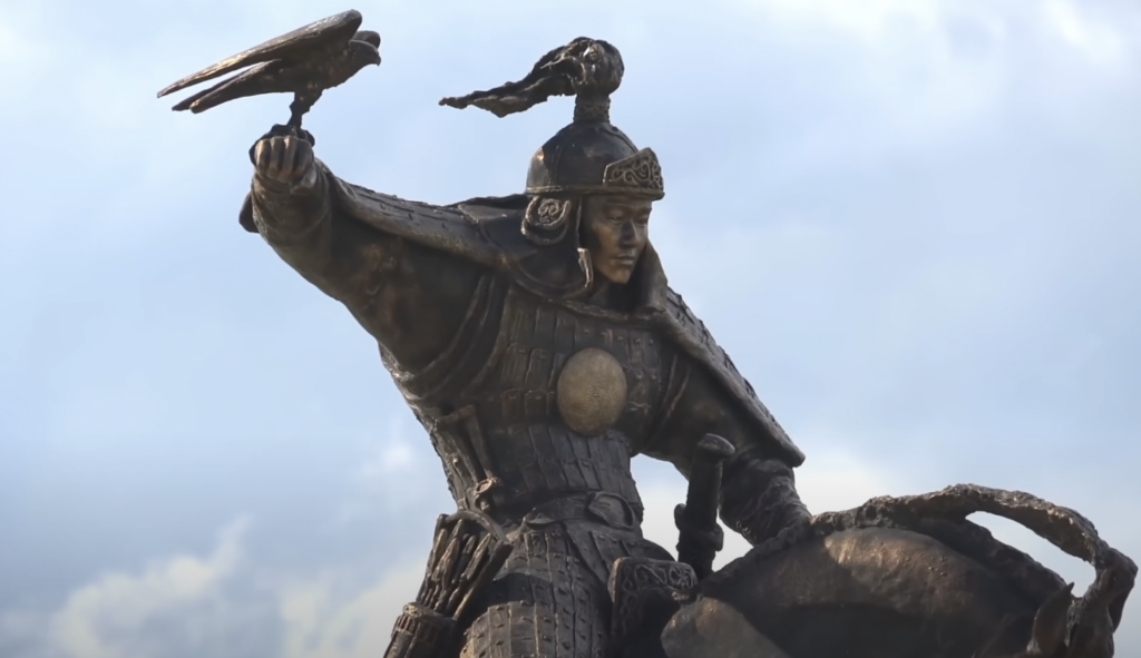 Statue of a warrior wearing ornate armor and a helmet, holding a falcon on the raised right arm. The warrior appears poised and fierce, set against a cloudy sky. The statue conveys a sense of strength and freedom.