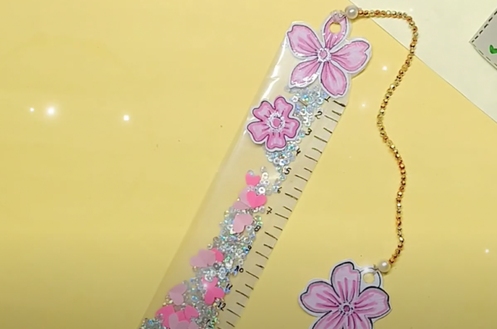 A decorative ruler featuring pink floral designs and glitter sequins inside a clear body. It has a gold chain with a pearl attached at the top. The ruler is placed on a light yellow surface.