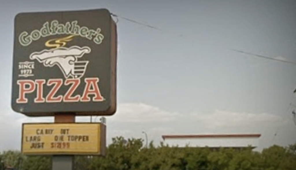 A tall sign displays "Godfather's Pizza" with a steam design. Beneath, a marquee advertises "Carry Out Large 1-Topper Just $7.99." The background shows a clear sky and some trees.