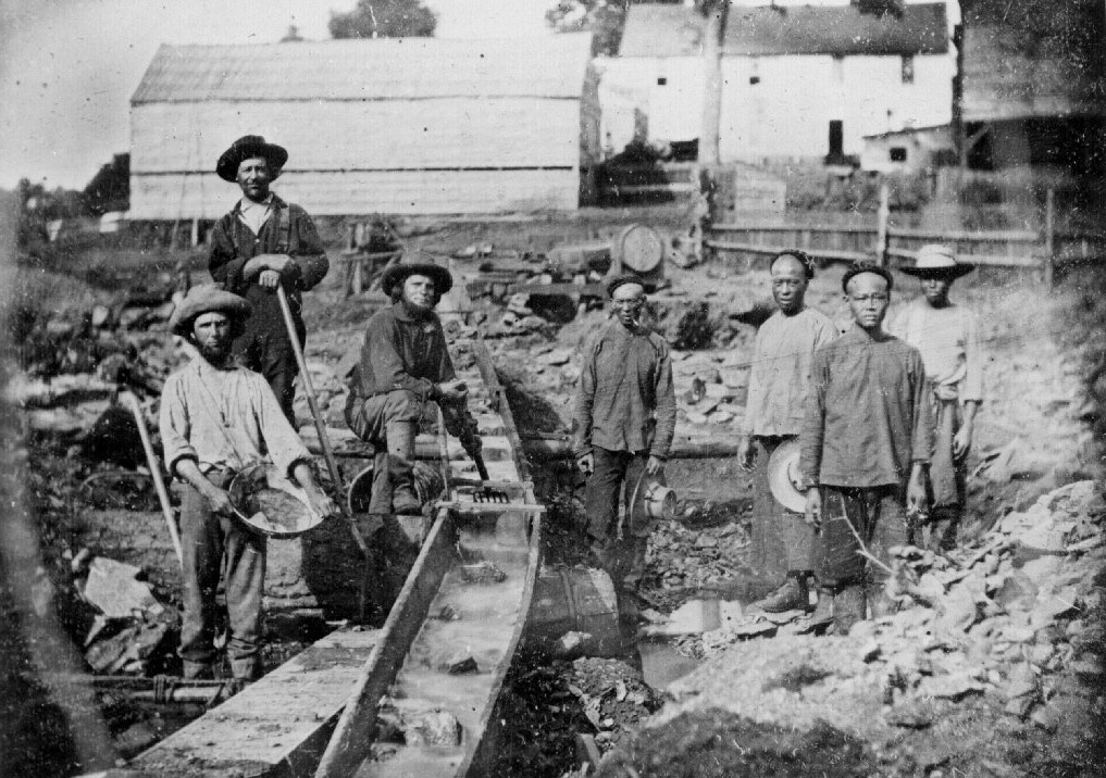 A historical black and white photo depicts a group of men in work attire standing beside a wooden sluice in a rocky area, likely a mining site. Buildings and equipment are visible in the background, indicating an industrial setting.