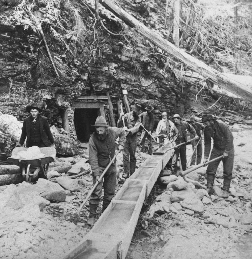 Old black and white photo of gold miners working with long wooden sluice boxes and shovels in a rocky area near a mine entrance. Several men are engaged in various tasks, surrounded by trees and equipment.