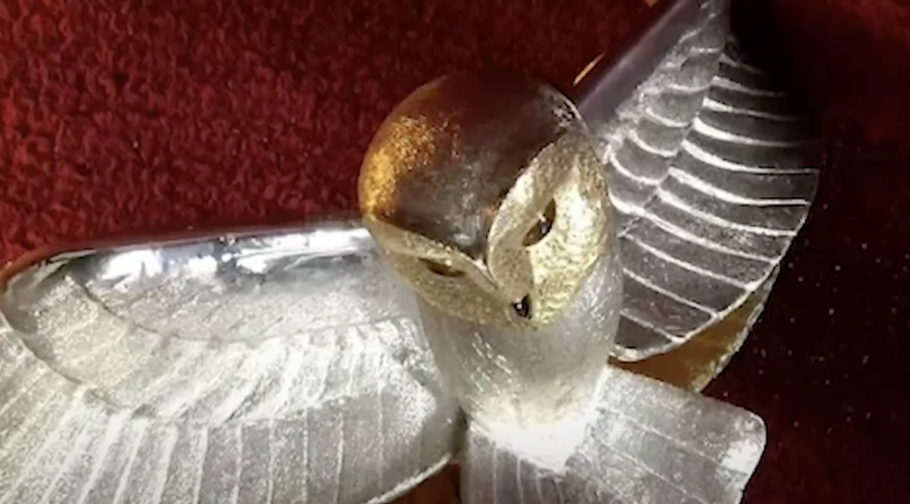 A silver and gold owl figurine with outstretched wings rests on a red textured surface. The owl's head is golden with distinct eyes, while the body and wings are silver with ribbed detailing.