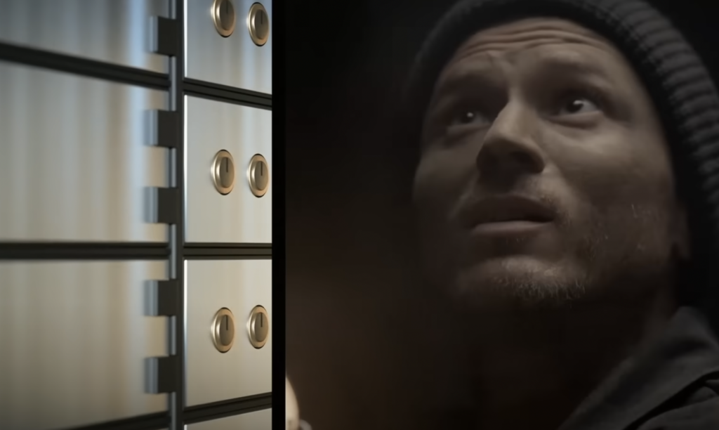 A man wearing a beanie and looking upwards stands in a dimly lit environment. On the left, there is a close-up of metallic safe deposit boxes with round handles.