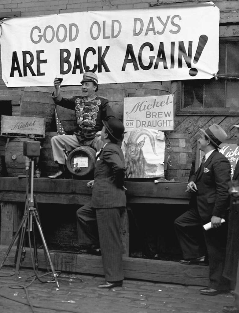 A man in a uniform enthusiastically raises a mug while standing on barrels with a sign reading "GOOD OLD DAYS ARE BACK AGAIN!" Two men in suits watch. There's a Michel beer poster and wooden crates around them, suggesting a celebration or event.