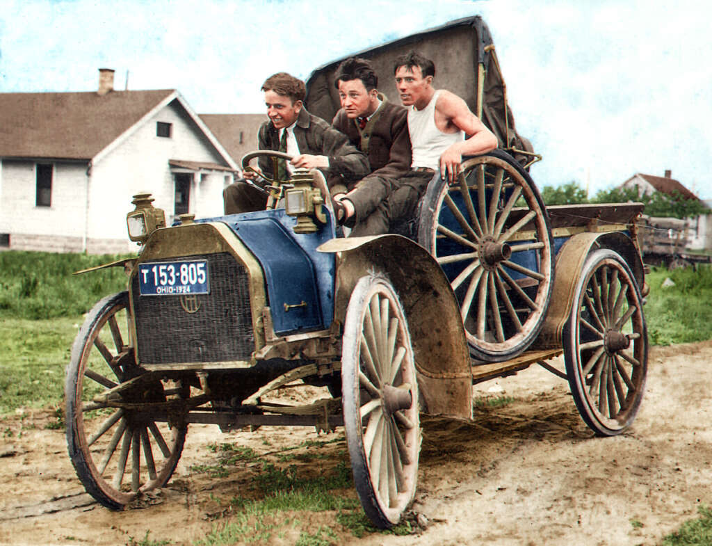 Three men are riding an early 20th-century open-top car on a dirt road. The car has wooden wheels and a blue body. The driver and one passenger wear suits, while the man in the back is in casual attire. A house is visible in the background.