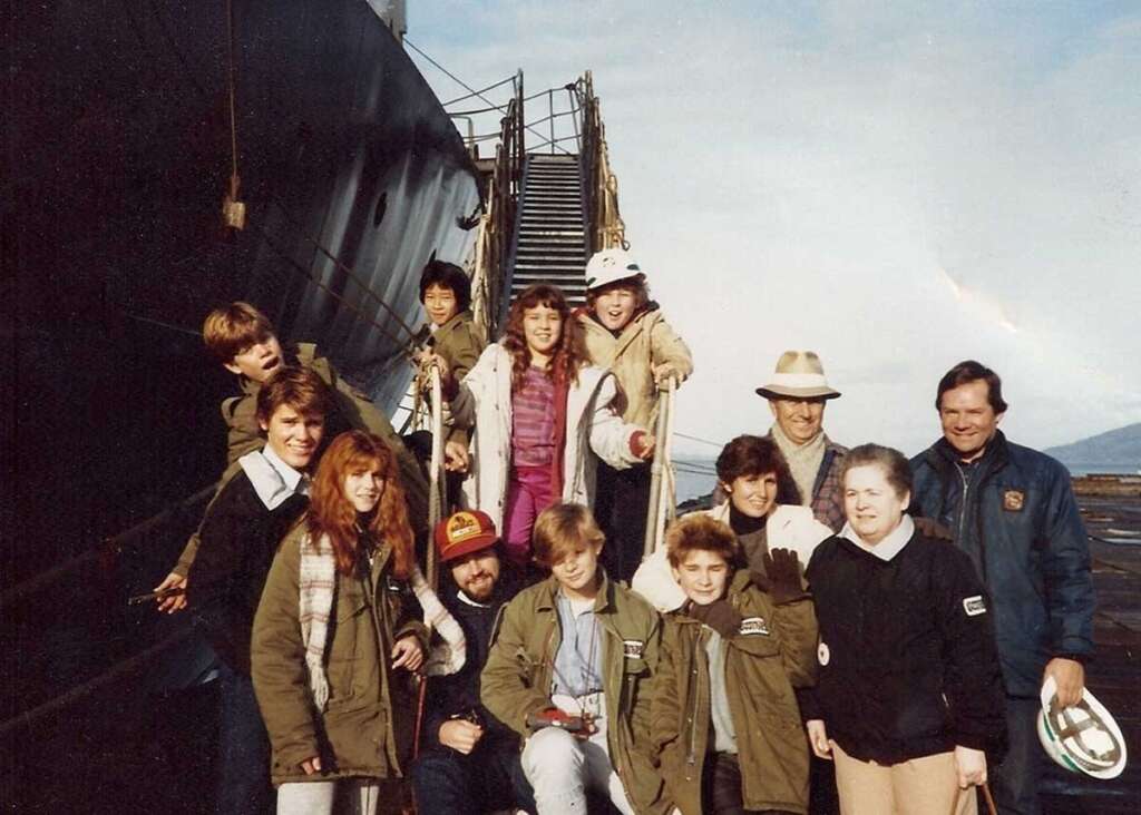 A group of twelve people, including children and adults, pose together outdoors in front of a large ship. They are dressed in casual, warm clothing. The sky is clear, and there is a visible staircase leading up to the ship in the background.