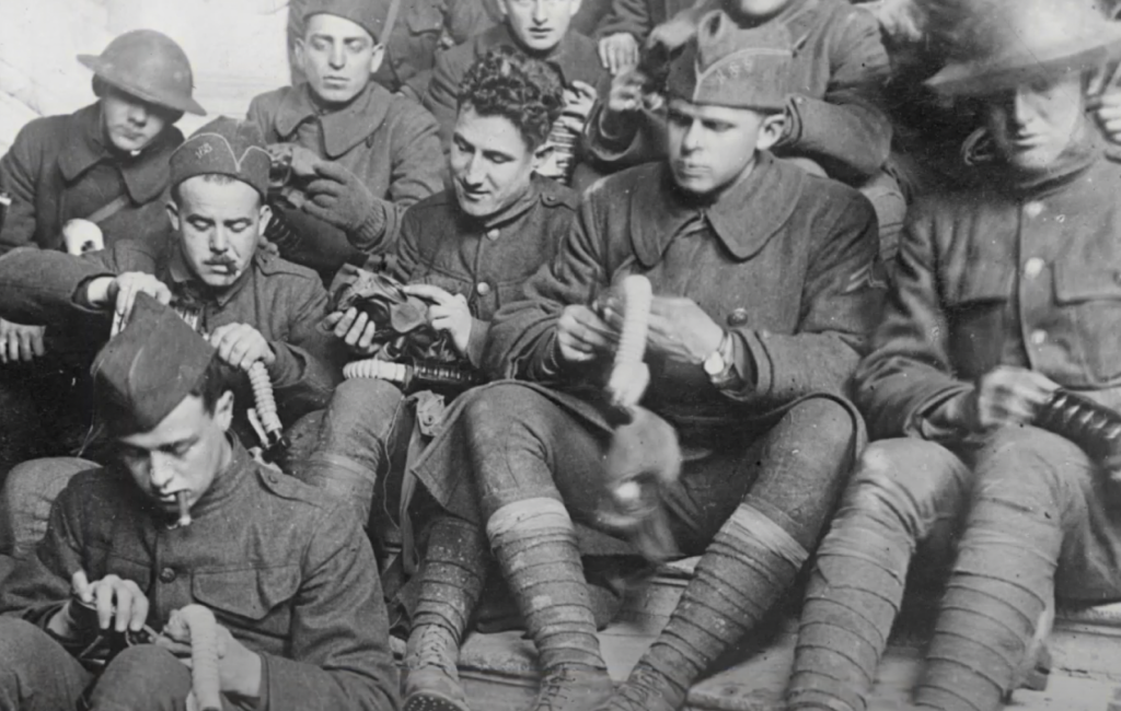 A group of soldiers in historical military uniforms are seated closely together, knitting. Most are focused on their knitting, holding yarn and needles. The scene evokes a sense of camaraderie among the soldiers.