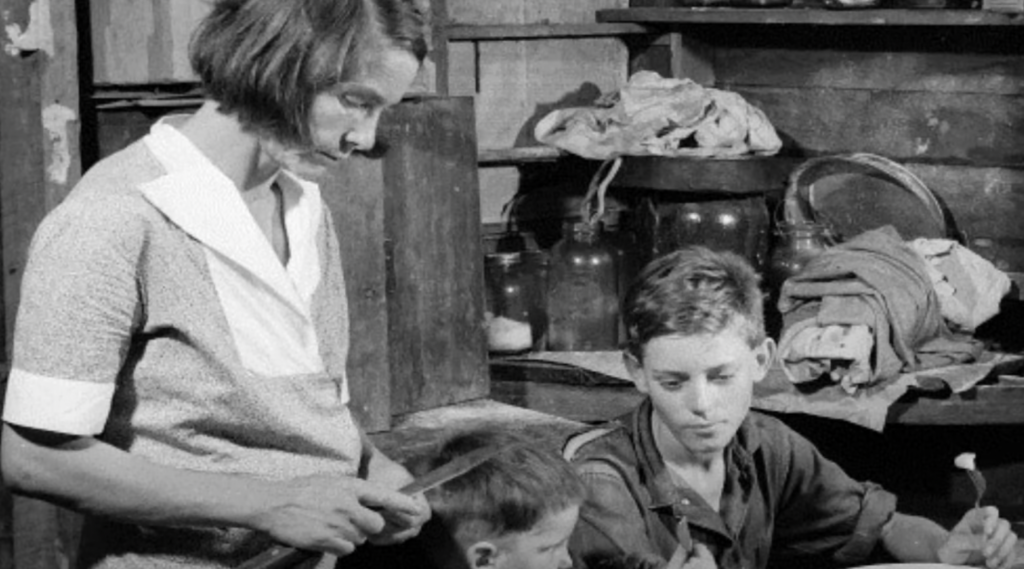 A woman stands slicing vegetables at a table with two young boys seated beside her. The room appears rustic, with shelves holding jars and cloth behind them. The boys are focused on tasks in front of them.