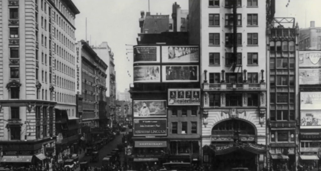 A vintage black-and-white photo of a bustling urban street. Tall buildings line both sides, with large billboards and advertisements covering some facades. Crowds and vehicles fill the road, depicting early 20th-century city life.