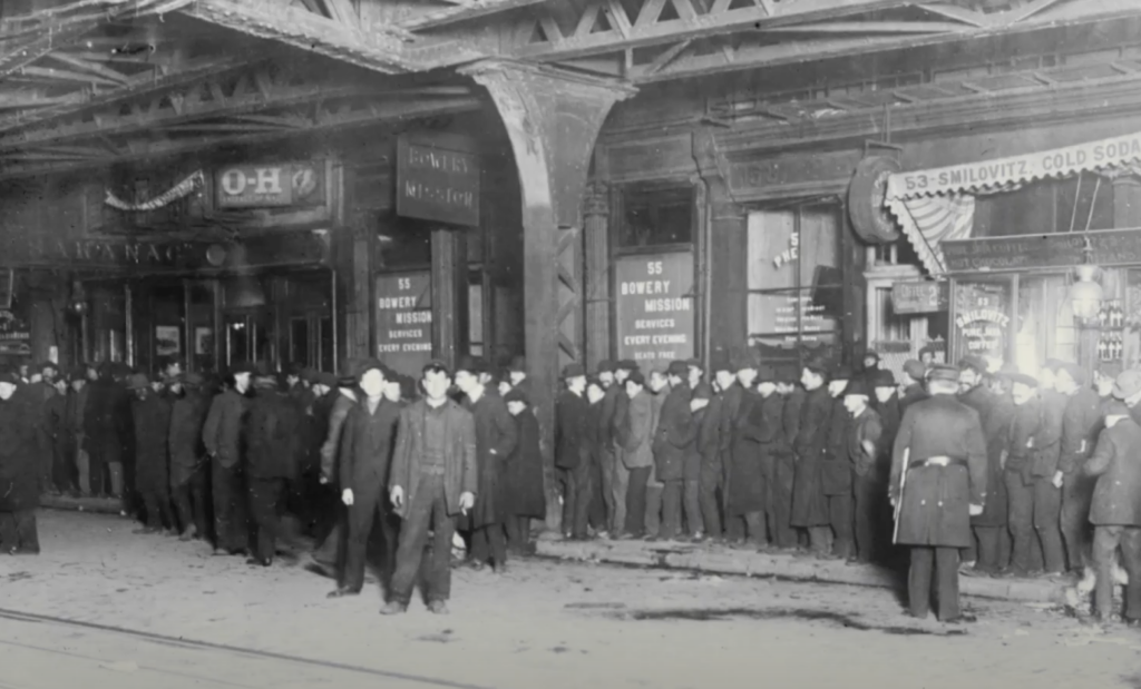 Black and white photo of a long line of men dressed in dark coats and hats, standing outside the Bowery Mission. The scene appears under an elevated railway in a city. Signage reads "Bowery Mission," with other shop signs visible.