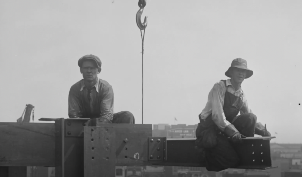 Two construction workers sit on a steel beam high above a city skyline. One man wears a flat cap and the other a wide-brimmed hat. A crane hook hangs between them. Both are dressed in work shirts and overalls. City buildings are visible in the background.