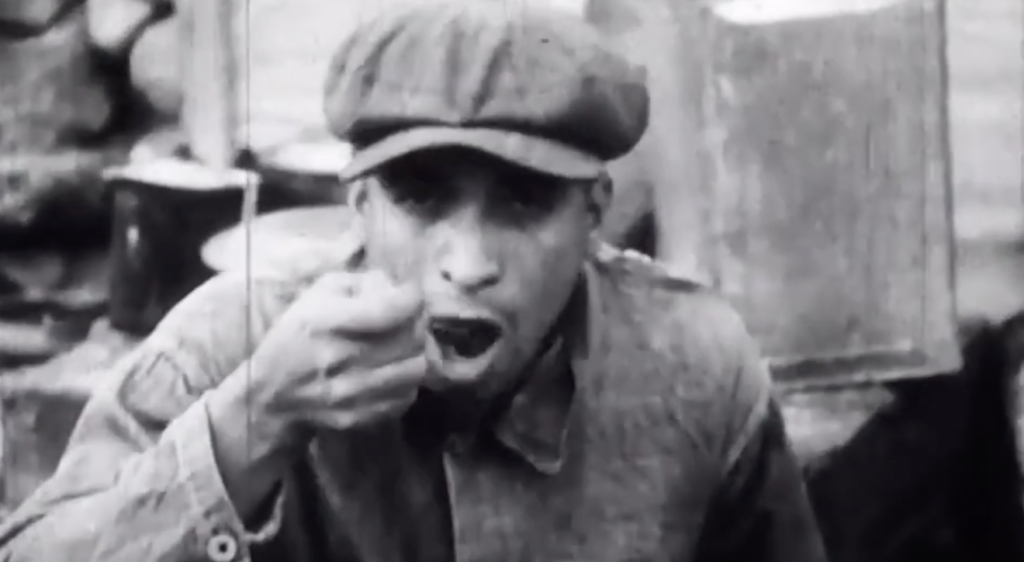 A black and white image of a person wearing a cap and eating with a spoon. They appear to be outdoors and are dressed in a jacket. Their facial expression suggests focus on the food. Background details are blurred.