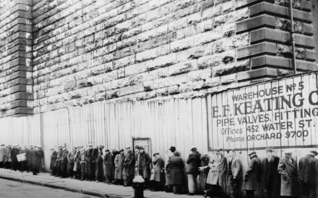 Black and white photo of a long line of people in coats and hats standing against a high brick wall. A sign reads "Warehouse No. 5, E.F. Keating Co., Pipe Valves, Fittings, Offices 452 Water St., Phone ORchard 9700.