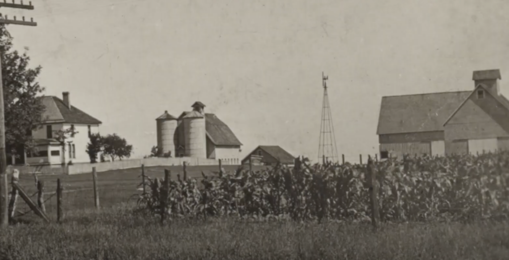 A vintage rural landscape featuring a farmhouse on the left, grain silos, a windmill, and a barn on the right. Cornfields are in the foreground, and a few trees dot the background under a clear sky.