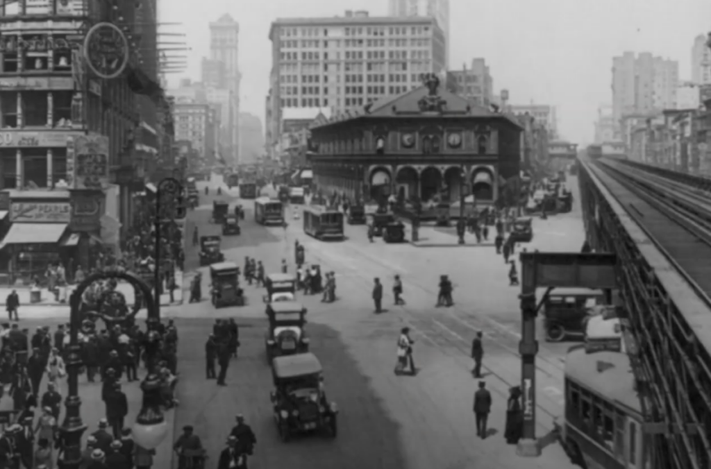 A busy city street scene from the early 20th century. People walk and cars drive on wide roads. A clock is on a building on the left, and a multi-story structure stands in the background. Elevated train tracks are visible on the right.