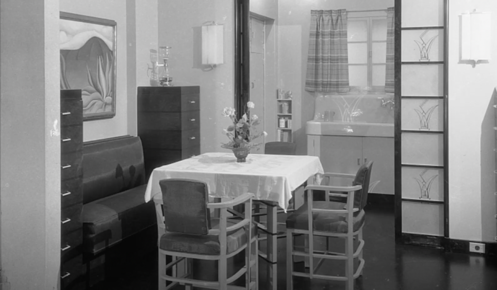 A vintage dining room with a square table covered by a white tablecloth, surrounded by four chairs. A cushioned bench and a tall cabinet are in the background. A curtained window and shelves are visible in a connected room. A vase with flowers is on the table.