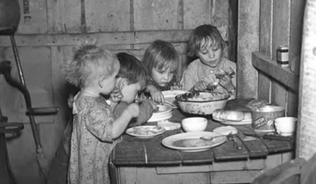 Four young children sit closely around a wooden table in a rustic room, eating from a large bowl and plates. The table holds a loaf of bread and cups. The background shows wooden walls and basic furnishings.