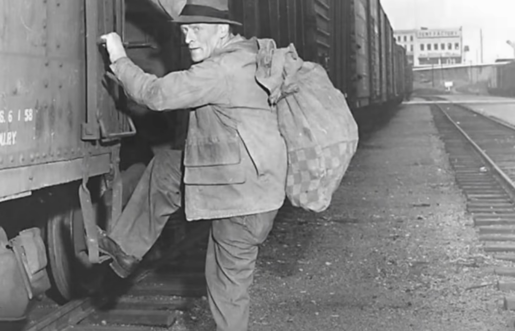 An older man wearing a hat and coat climbs into a freight train car, holding a large, checkered sack over his shoulder. The train is stationary on a railway track, and industrial buildings are visible in the background.