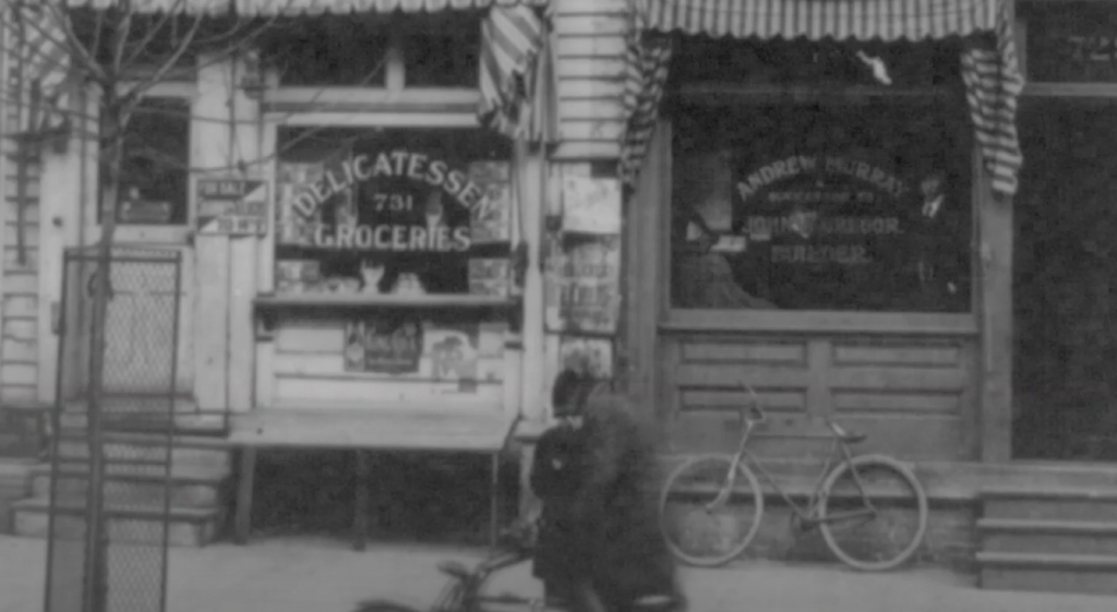 Black and white photo of a storefront with "Delicatessen Groceries" on the window. A bicycle leans against the building. Striped awnings hang above, and a child is seen in motion on a tricycle in the foreground.