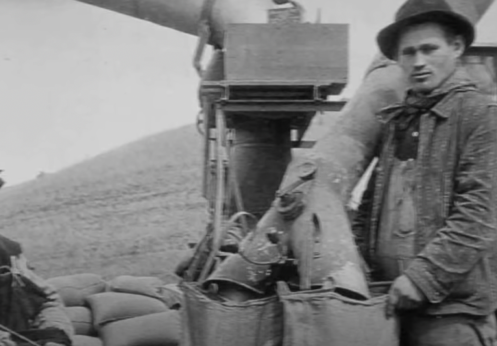 Black and white photo of a man standing next to a machine filling sacks with grain. He is wearing a hat and jacket, looking towards the camera. The background shows a slight hill and more sacks stacked around.