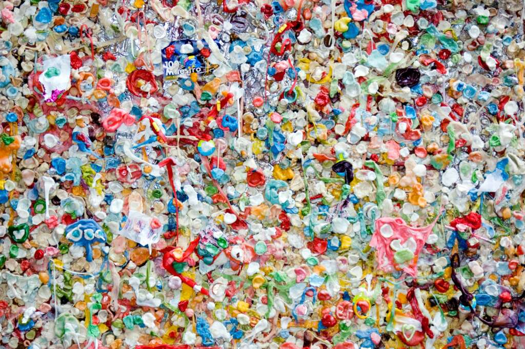 A colorful wall covered in countless pieces of chewed gum, creating a vibrant and chaotic mosaic of various sizes and colors. Mixed amongst the gum are occasional pieces of paper and other small objects.