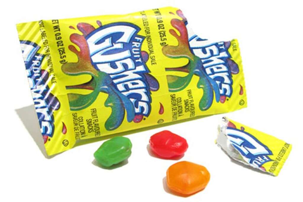 A partially opened package of Fruit Gushers, featuring a colorful design, lies on a white surface. Three Gushers candies—one red, one orange, and one green—are scattered beside the package.