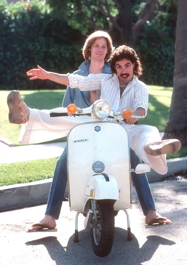 Two people joyfully pose on a white Vespa scooter. The person in front has a handlebar mustache, and the person behind has long hair. Both are wearing casual summer clothing and sandals, with a lush garden visible in the background.