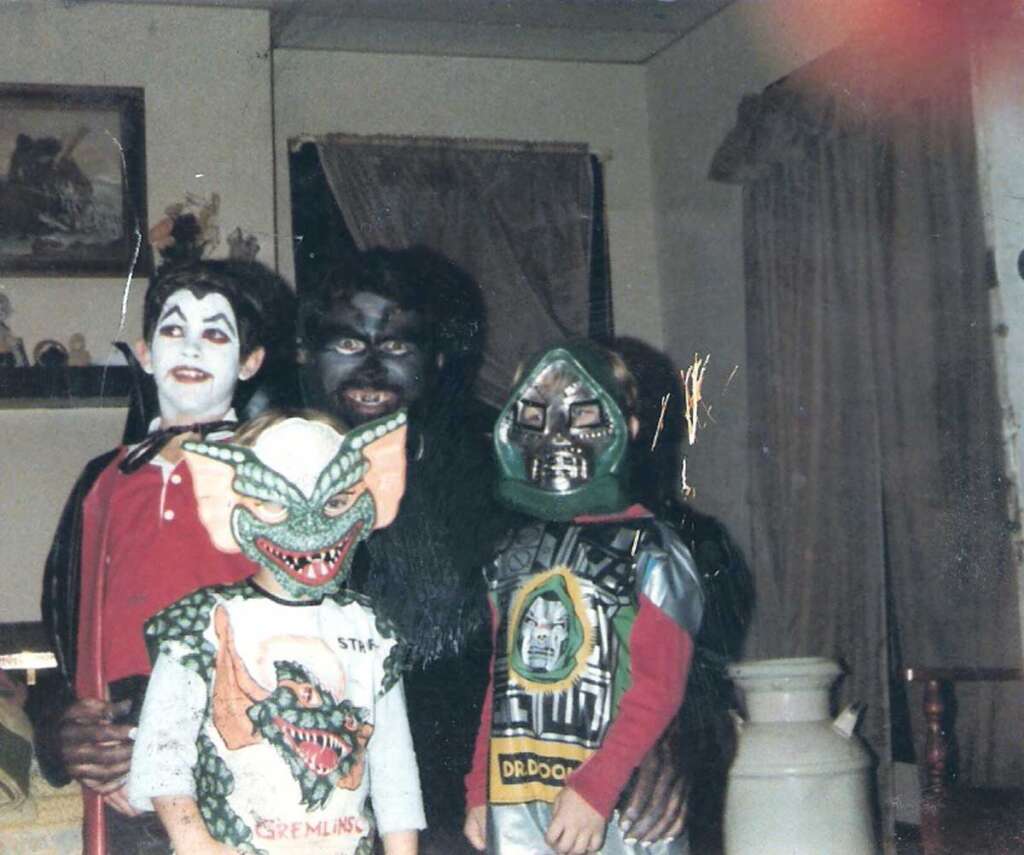 Four children in Halloween costumes pose in a living room. The costumes include a vampire, a gremlin, a werewolf-like creature, and a robot. The room has framed pictures and curtains in the background.