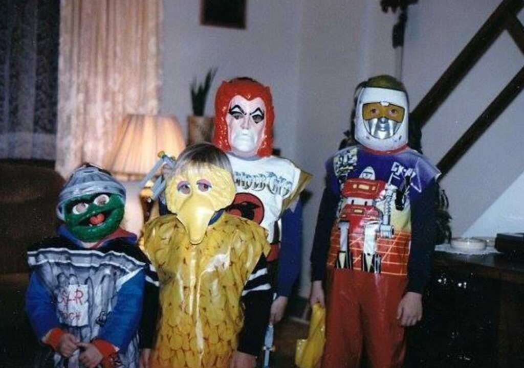 Four children in a living room are wearing colorful Halloween costumes and masks. The costumes are a mix of a green character, a yellow bird, a figure with a red mask, and a robot-like character. A lamp and curtains are in the background.