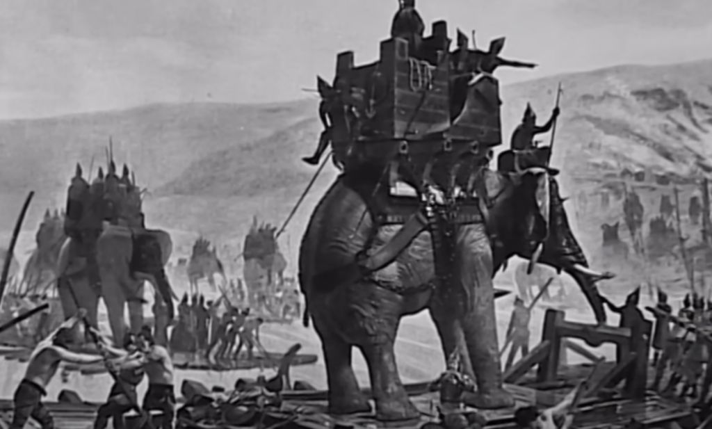 A historical illustration depicts a battle scene with armored elephants and warriors. Soldiers ride the elephants, wielding spears, as others engage in combat on the ground. The background features hills and scattered troops.