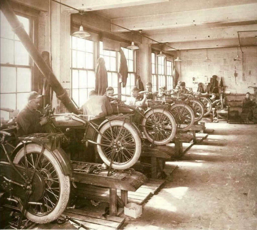 A sepia-toned image shows several workers assembling motorcycles in a row inside a factory. Each worker focuses on a motorcycle, with tools and parts visible on tables. Large windows provide natural light to the spacious workshop.