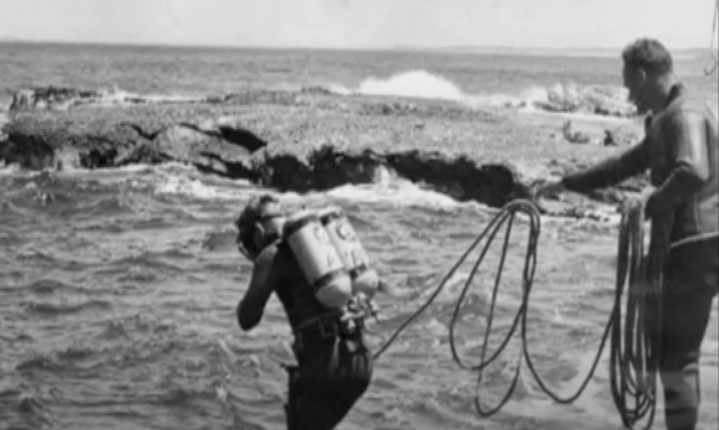 A vintage black-and-white photo shows a person in scuba gear preparing to dive into the ocean, holding onto a rope. Another person stands on the rocky shore, assisting by holding the rope. Waves crash in the background.