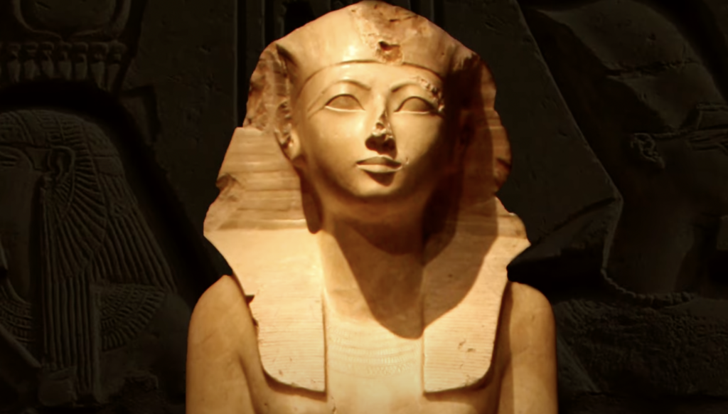 A limestone bust of an Egyptian pharaoh, adorned with a nemes headdress. The sculpture is set against a dark background featuring carvings with hieroglyphics and figures, highlighting the bust's details and serene expression.