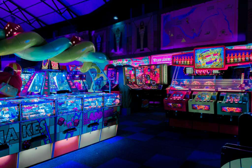 A dimly lit arcade filled with colorful neon lights reflects off various gaming machines. The vibrant displays feature games like slot machines and pinball, creating a lively and energetic atmosphere.