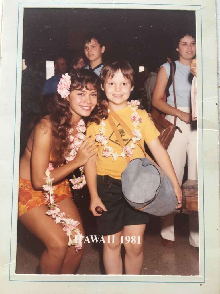 A woman in a floral lei and orange outfit smiles and poses with a young child wearing a lei and yellow shirt. The child holds a large bag. The image is labeled "Hawaii 1981." Several people are in the background.